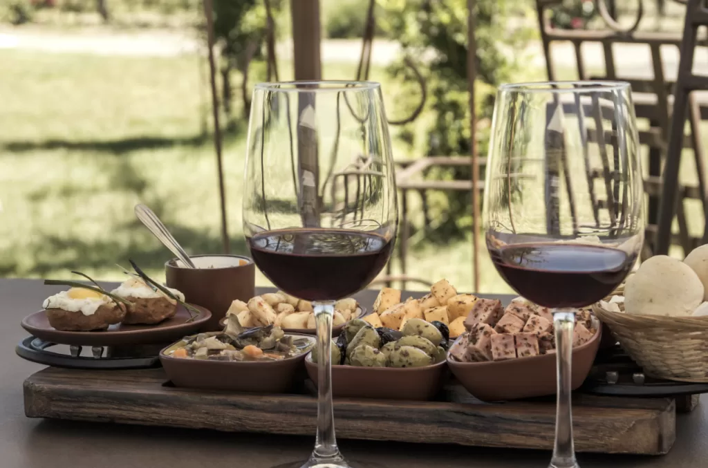 napa valley outfitTwo glasses of red wine are placed in the foreground with a wooden board featuring a variety of cheeses, olives, and other snacks set against a lush vineyard background.